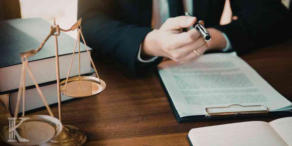 Prawnik w garniturze trzymający pióro nad dokumentem umieszczonym w podkładce z klipsem; po lewej stronie na grubych księgach stoi mosiężna waga szali.