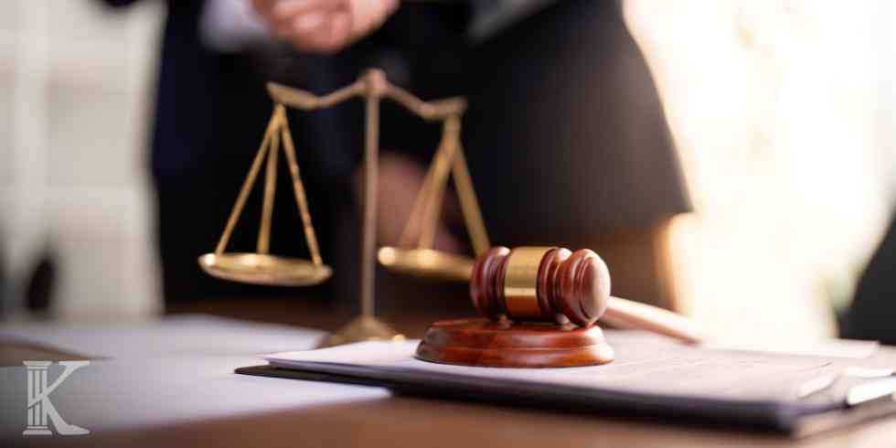 Młotek sędziowski na podstawce oraz złota waga sprawiedliwości stojące na biurku z dokumentami; w tle rozmyta postać prawnika.