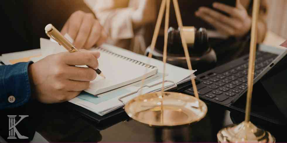 Close-up of a person writing in a notebook at a desk with scales of justice and a laptop, symbolizing law practice.