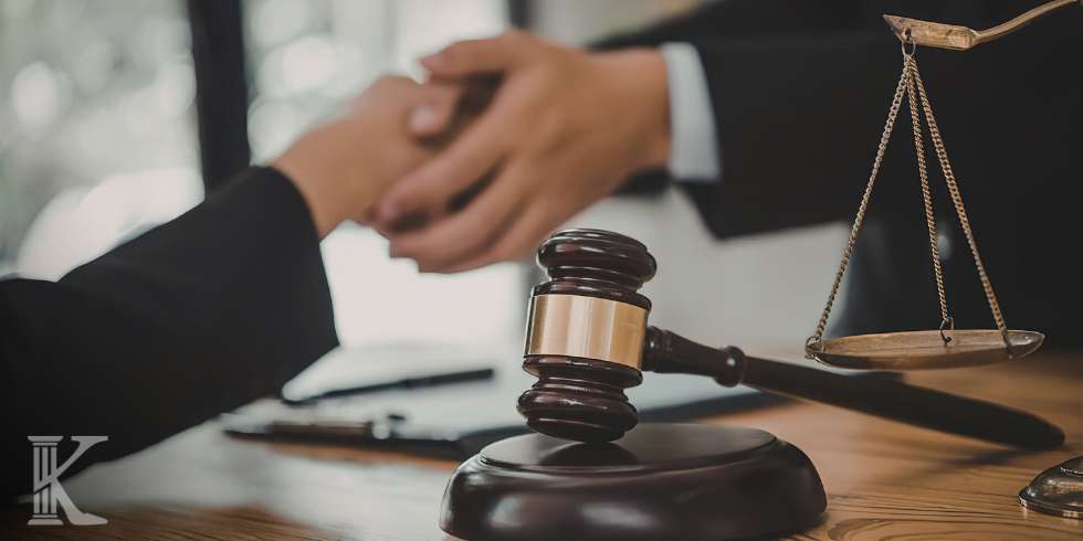 Młotek sędziowski na podstawce oraz złota waga sprawiedliwości stojące na biurku z dokumentami; w tle rozmyta postać prawnika.