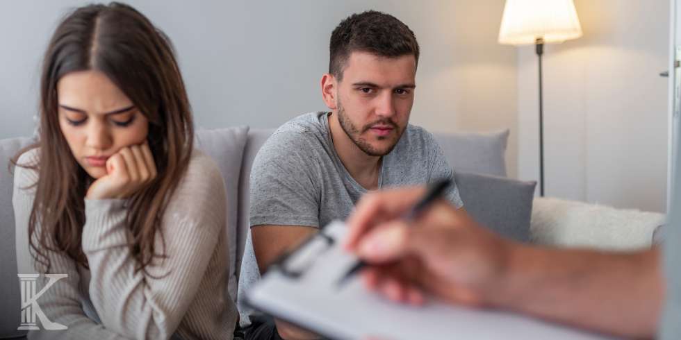 Couple sits on a sofa in a counseling session; the woman looks upset, the man appears concerned as a counselor takes notes.