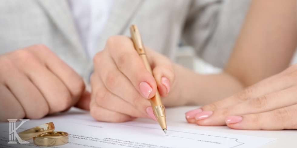 Close-up of hands signing a document with a gold pen, pink nails visible, rings on the desk nearby.