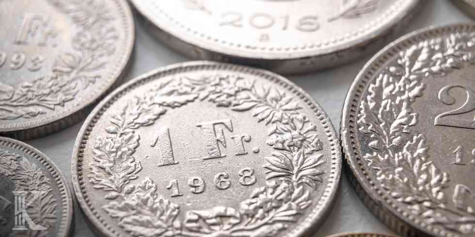 Makrofotografia srebrnych monet franka szwajcarskiego; na pierwszym planie moneta o nominale 1 frank z datą 1968 w otoczeniu roślinnego wieńca.