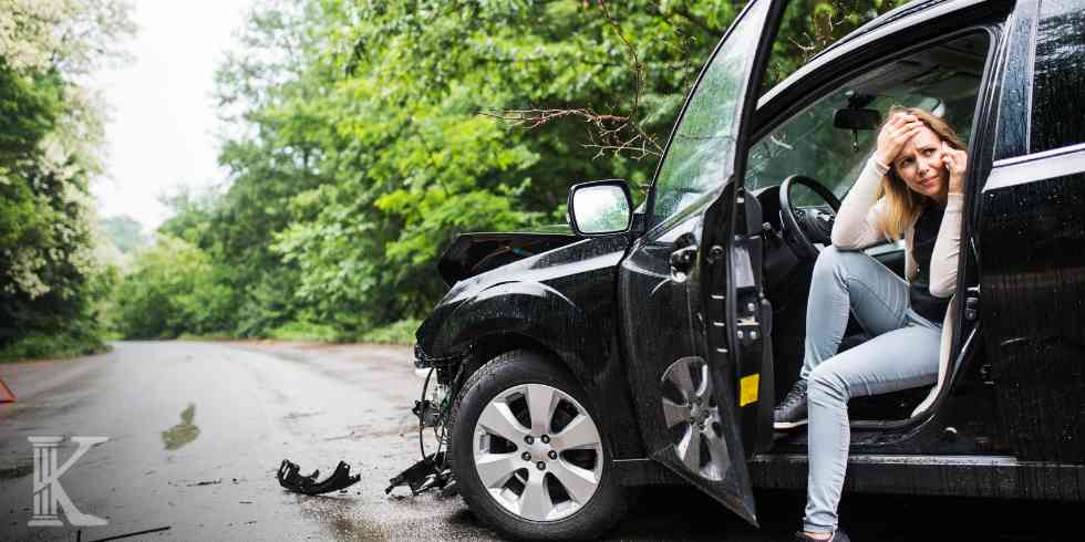 Rozbite czarne auto na leśnej drodze po wypadku, obok siedząca w otwartych drzwiach kobieta rozmawiająca przez telefon i trzymająca się za głowę