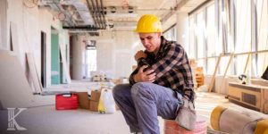 Mężczyzna w żółtym kasku ochronnym i koszuli w kratę siedzący na placu budowy wewnątrz budynku, trzymający się za obolały łokieć z grymasem bólu na twarzy.