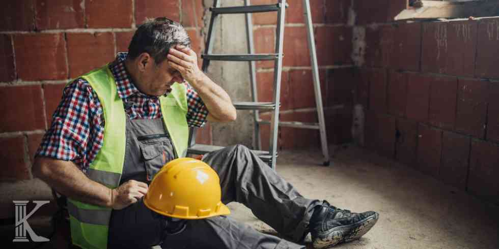 Starszy mężczyzna w kamizelce odblaskowej i roboczych ogrodniczkach siedzący na ziemi obok aluminiowej drabiny na placu budowy. Mężczyzna trzyma się za czoło, obok leży jego żółty kask ochronny.