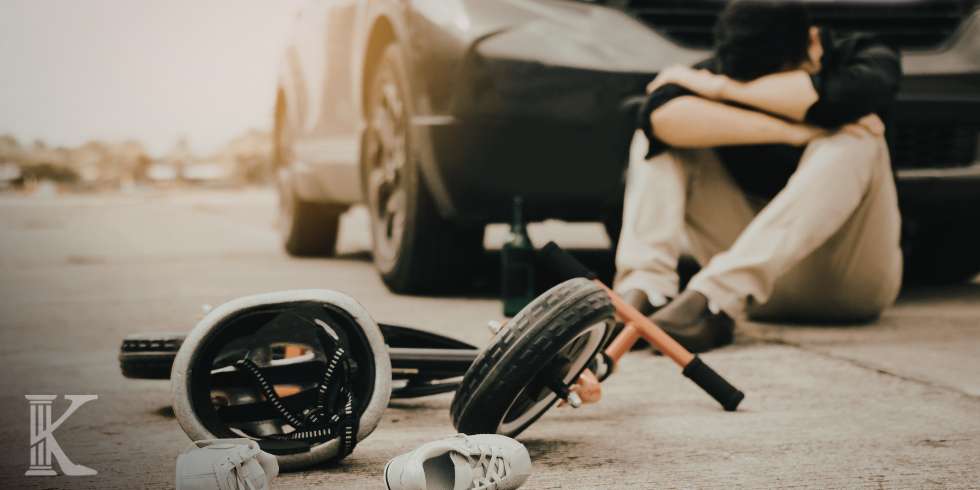 Person sits on the pavement next to a car after a bicycle crash, with a helmet, wheel, and tools scattered nearby on the ground.