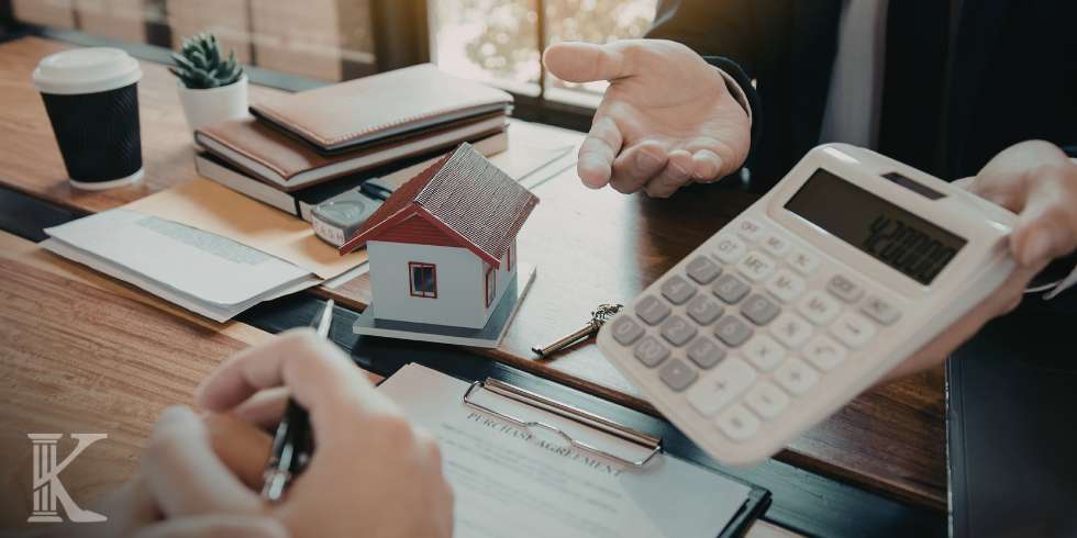 Person gesturing toward a miniature house on a desk during a mortgage discussion, with a calculator, papers, and coffee nearby.