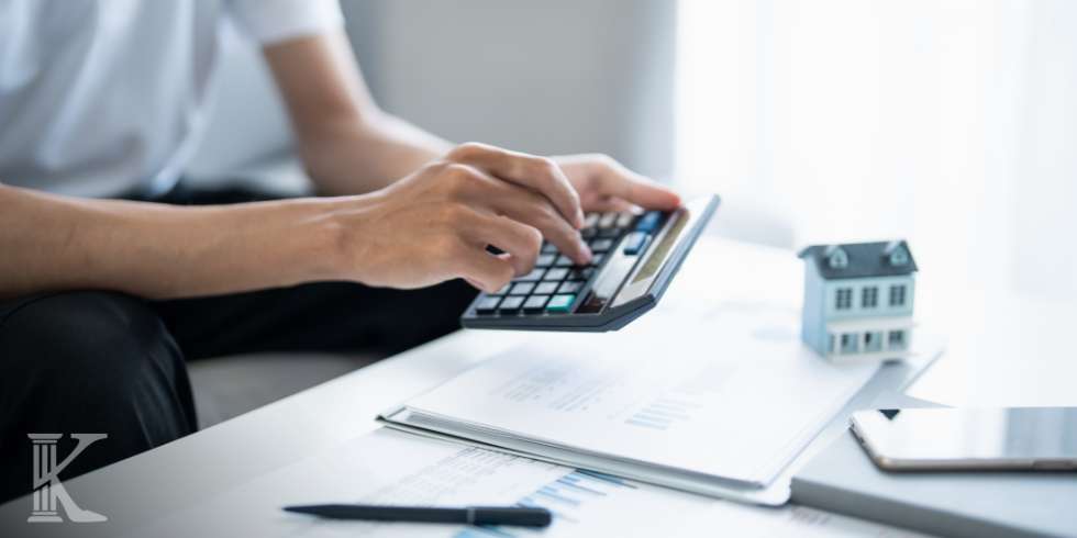 Person uses a calculator over real estate documents on a bright desk, with a small house model nearby