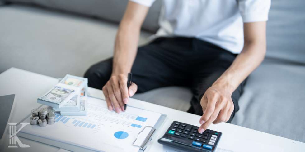 Person calculating finances: stacks of cash and coins on a table, with charts on a clipboard and a calculator nearby.