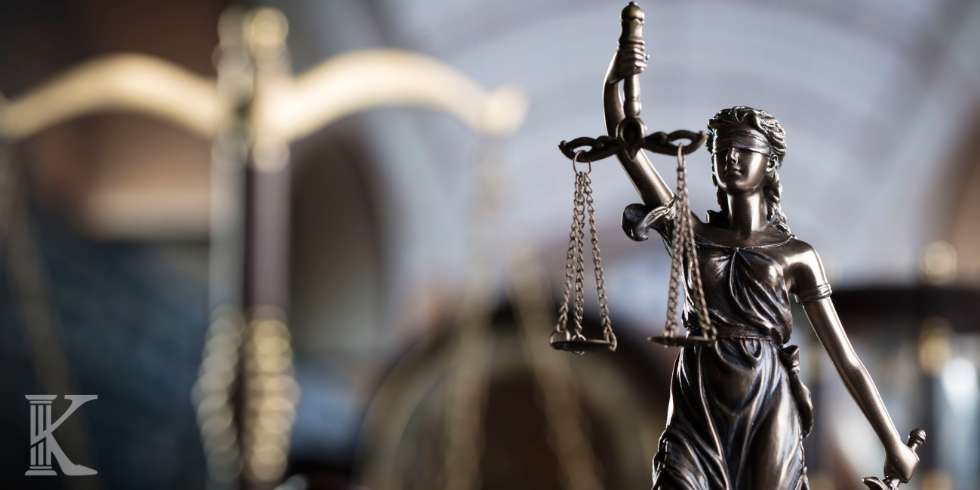 Bronze statue of Lady Justice holding scales, with a blurred courtroom background.