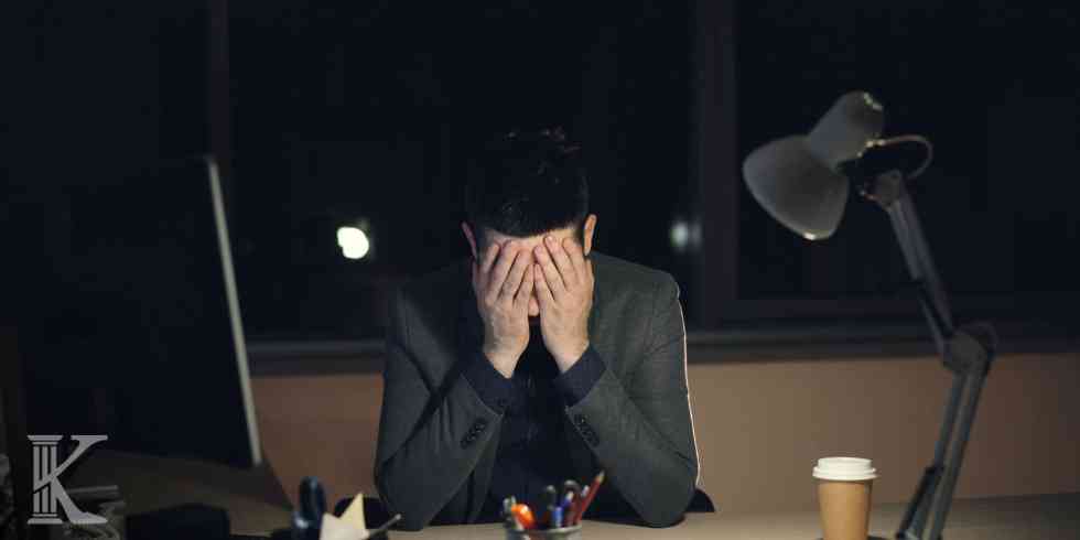 A man in a suit sits at a desk at night, head in his hands showing stress, with a desk lamp and coffee cup nearby.