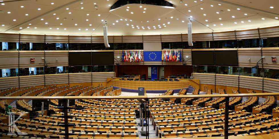 A wide view of a large, empty European Parliament chamber with curved rows of seats, desks, and a central podium, EU flags in the back.