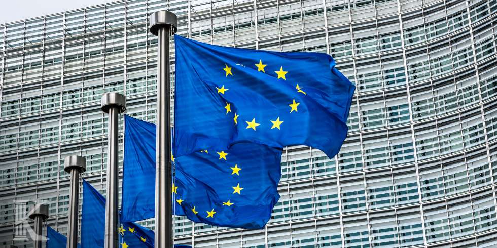 EU flags wave outside a modern glass building, symbolizing the European Union hub or headquarters.