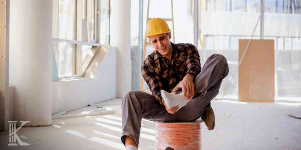 Construction worker in a yellow hard hat sitting on a coiled rope, examining rolled blueprints in an unfinished indoor space with large windows and scattered tools.