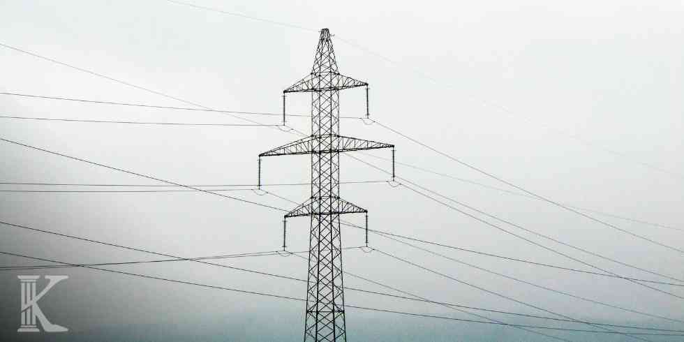 High-voltage transmission tower with multiple power lines extending across a pale, cloudy sky in a rural setting.