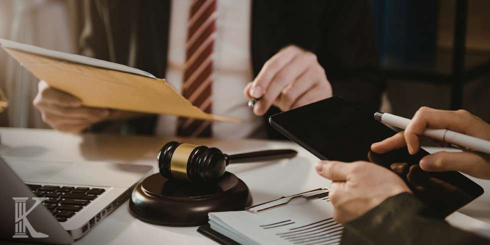 Gavel on a desk beside a laptop and documents, with hands using a tablet and pen in a legal office setting.