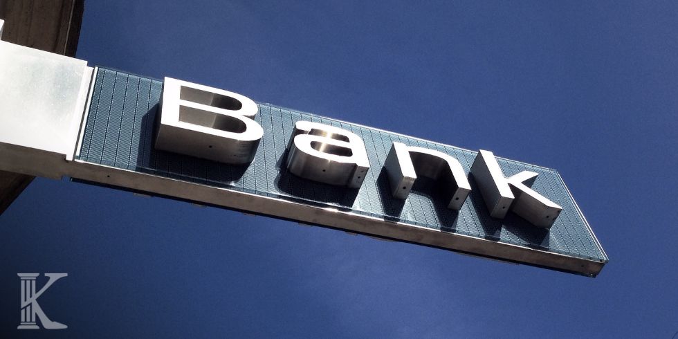 Slanted bank sign on a blue panel reading 'Bank' against a clear blue sky with a small logo in the corner.