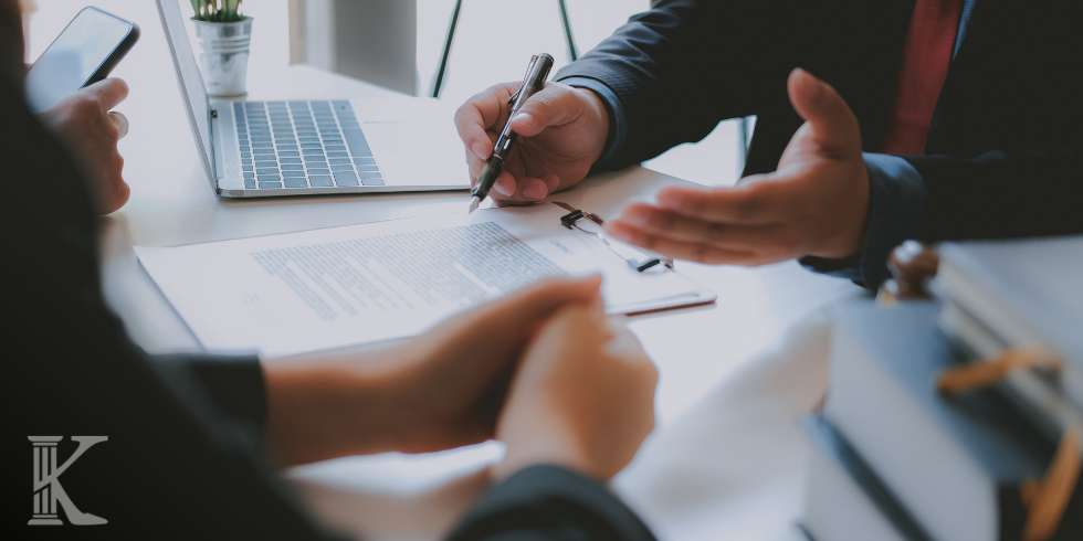Two professionals discuss a document at a desk with a laptop and pen in a business meeting.
