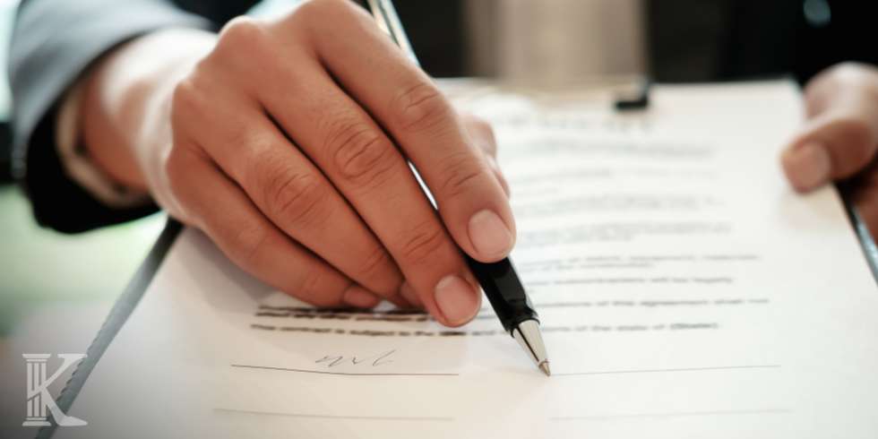 Close-up of a hand signing a document with a black pen on a clipboard or desk surface.