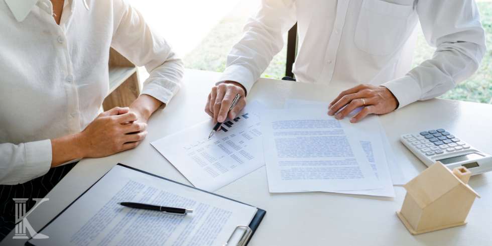 Two professionals in white shirts review documents and charts at a desk, with a pen, calculator, and a small wooden house model nearby.