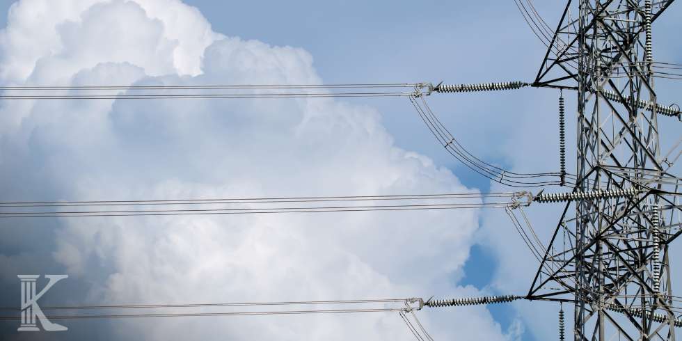 Electric power lines stretch across the sky with a lattice transmission tower on the right against a cloudy blue sky.