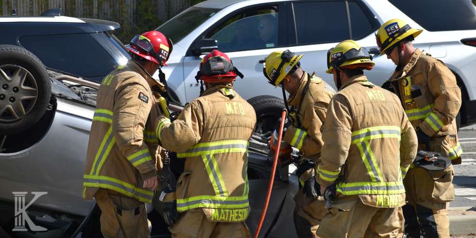 Firefighters in turnout gear work around an overturned silver car, using rescue tools at a crash scene.