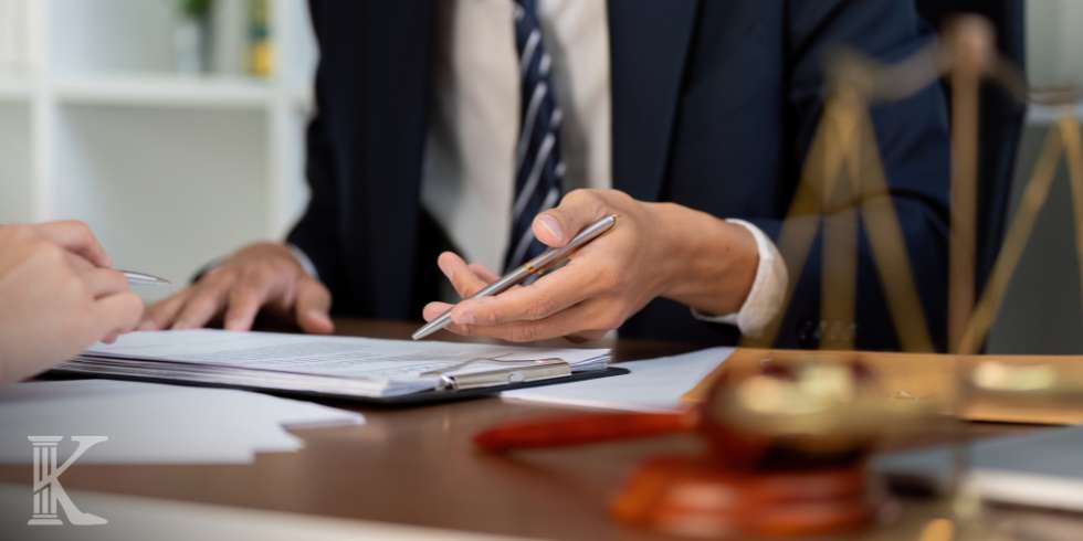 Lawyer in a suit signs documents at a desk, with a gavel in the foreground and scales of justice nearby.