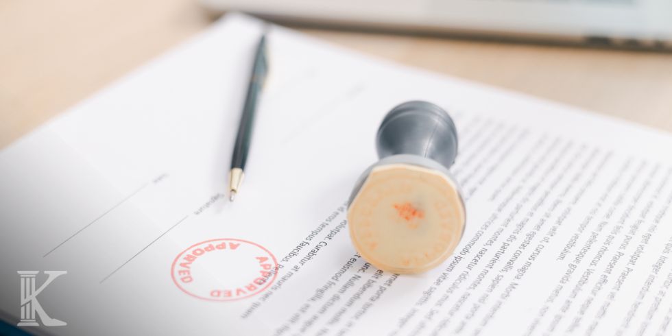 Document with a red "APPROVED" stamp, a black pen, and a rubber stamp resting on the page. Synoptic view of an approved contract or form.