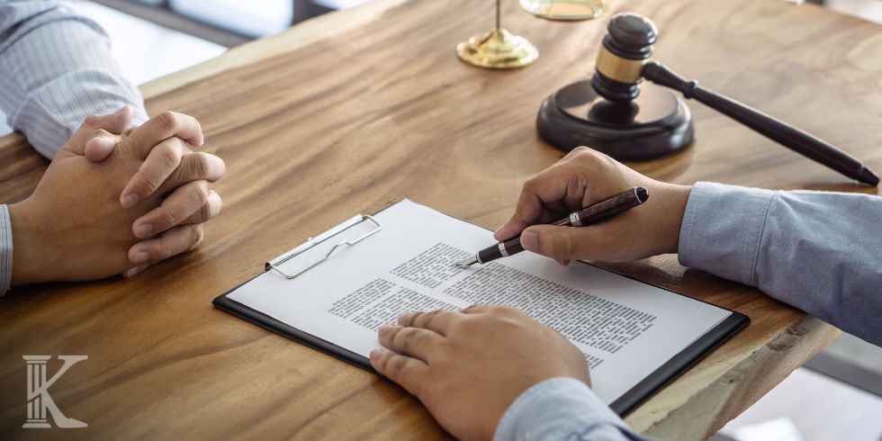 Legal signing ceremony: person signs a document with a pen while another person sits with clasped hands at a wooden table; gavel in the background.