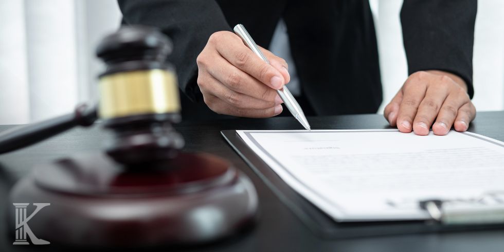 Hand signing a document at a desk with a gavel in the foreground, symbolizing a legal proceeding