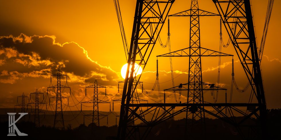 Silhouetted high-voltage transmission towers and power lines against an orange sunset sky on a flat horizon.