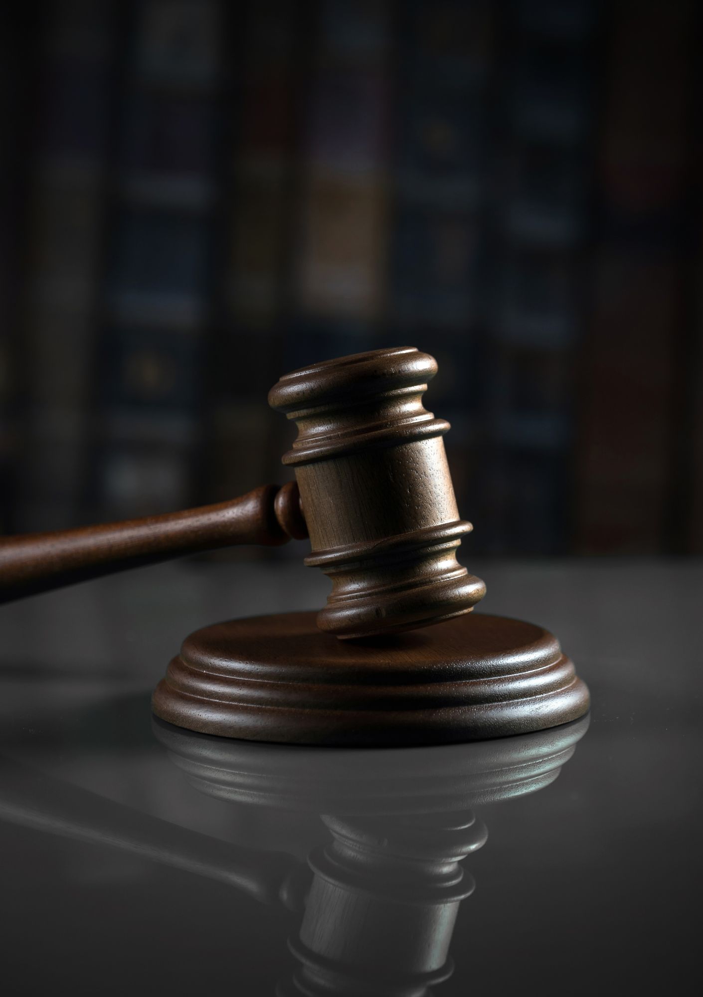 Close-up of a wooden judge's gavel resting on a round sound block, with a dark, blurred bookshelf background and its reflection on a glossy surface.