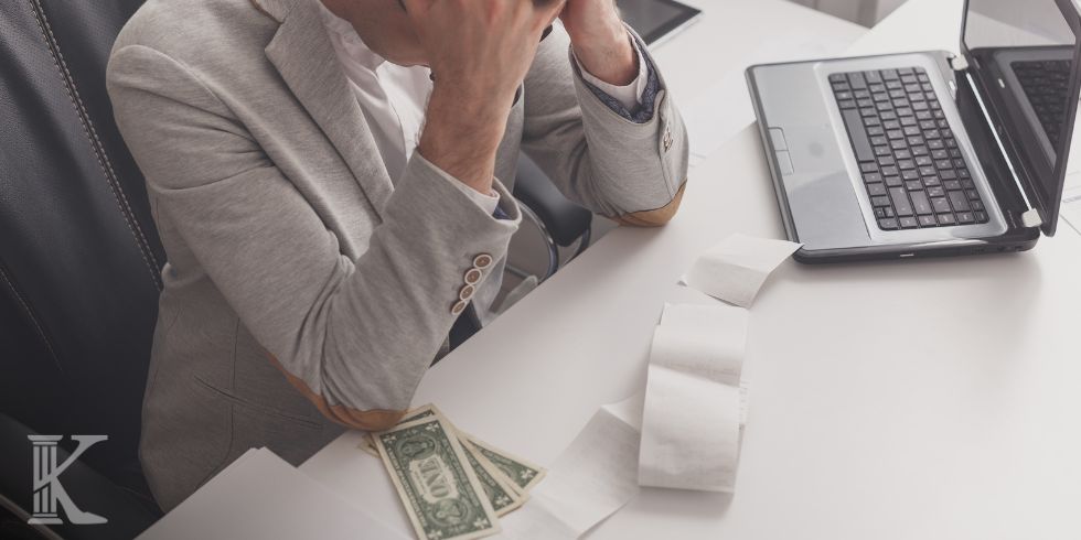 Osoba siedzi przy biurku, na którym leżą otwarty laptop, paragon i banknoty